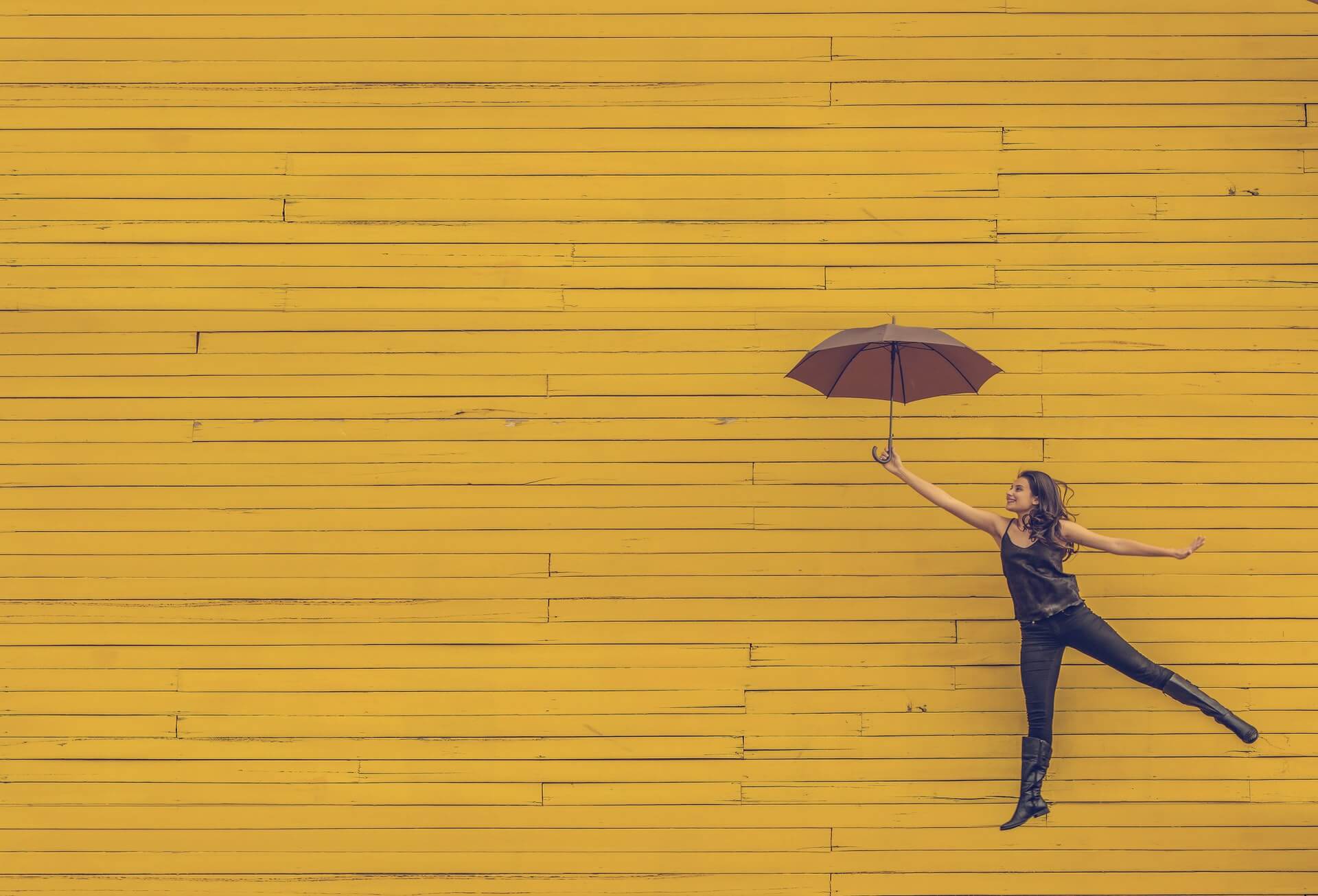 Żółta ściana. Na jej tle wesoła kobieta skacze z parasolką